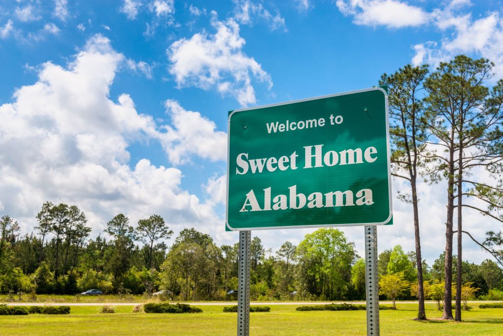Big green sign that says "Welcome to Sweet Home Alabama" in front of a blue sky and forrest