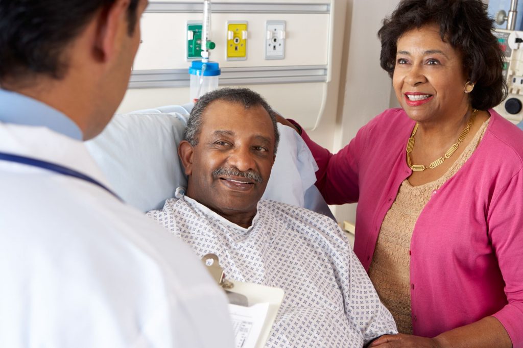 Black senior man in hospital bed with wife by his side talking with a doctor